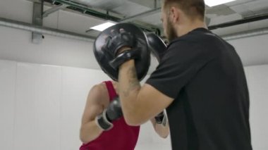 Beyaz odadaki bir boks antrenmanında, bir adam eldivenli ellerini koçun patilerine vurur. Darbelerden sapma ve dalış gerçekleştirin. Yavaş çekim.