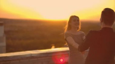 Aşık bir adam ve bir kadın gün batımında bir binanın çatısında birbirlerine bakarak dans ediyorlar.