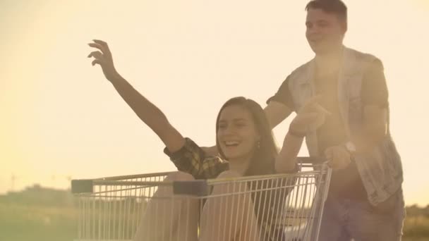Lens flare: Cheerful people couple man and woman at sunset ride supermarket trolleys in slow motion — Stock Video