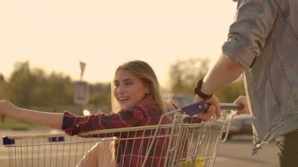 若いスタイリッシュなクーペは、スーパーマーケットの近くの屋外駐車場でショッピングカートで楽しい乗馬を持っています. — ストック動画