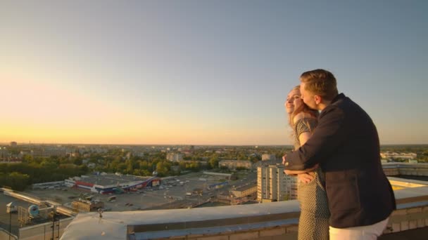 Un hombre y una mujer de pie en el techo al atardecer abrazan y miran la hermosa vista — Vídeos de Stock