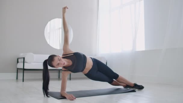 De brunette in het appartement doet een oefening plank terwijl staan in een statische positie op de laterale spieren van de buikspieren en het verplaatsen van haar hand op en neer. — Stockvideo