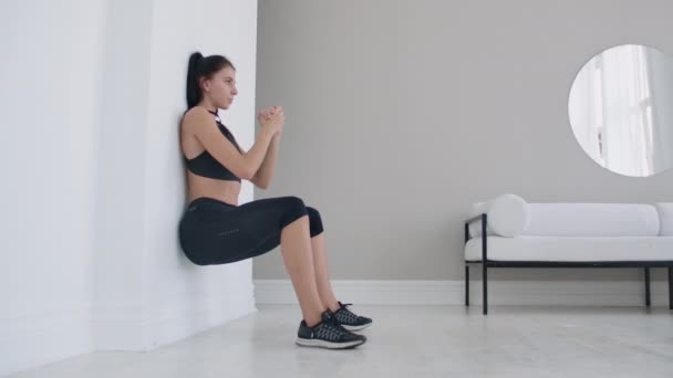 Uma mulher em uma posição estática realiza um exercício extenuante encostado a uma parede em uma posição sentada. Treinamento de resistência — Vídeo de Stock