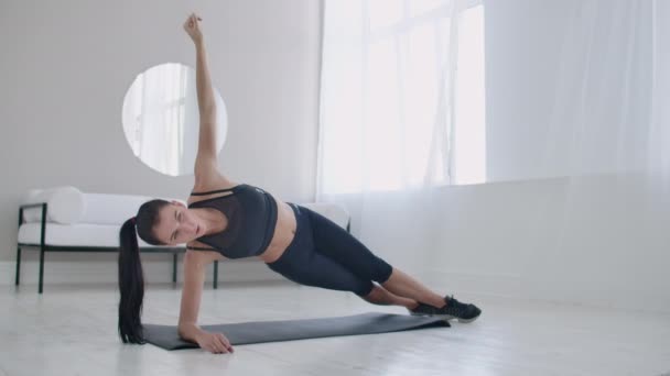 Brunetka w mieszkaniu robi deskę do ćwiczeń stojąc w statycznej pozycji na bocznych mięśniach brzucha i poruszając ręką w górę iw dół. — Wideo stockowe