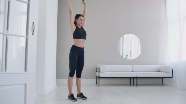 Otthoni gyakorlatok egy gyönyörű testnek, egy fiatal barna nő lassan felemeli a súlyzókat a karjaiban. — Stock videók