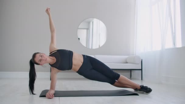 A morena no apartamento faz uma prancha de exercício enquanto está em uma posição estática nos músculos laterais dos abdominais e movendo a mão para cima e para baixo . — Vídeo de Stock