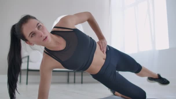 De brunette in het appartement doet een oefening laterale plank staan op haar knie en het verhogen van haar been omhoog. Fitnessmarathon — Stockvideo