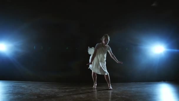 Una joven bailarina descalza salta al escenario y se mueve en cámara lenta con un vestido blanco suelto — Vídeo de stock