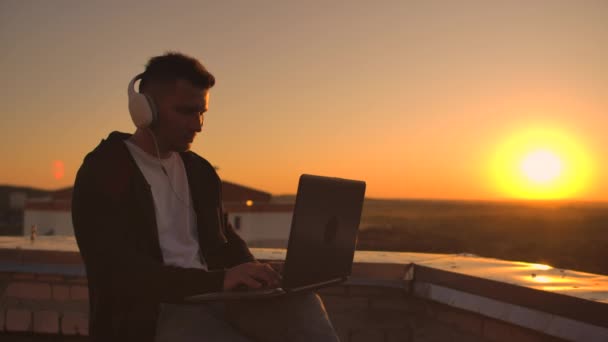 Vista traseira de um homem em fones de ouvido ouvindo música e trabalhando no telhado de um edifício ao pôr do sol com vista para a cidade a partir de uma altura. Telhado de um arranha-céu ao pôr-do-sol — Vídeo de Stock