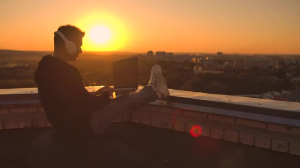 Un hombre en una sudadera con capucha se sienta en el techo y escucha música con auriculares escribiendo en un teclado portátil . — Vídeos de Stock
