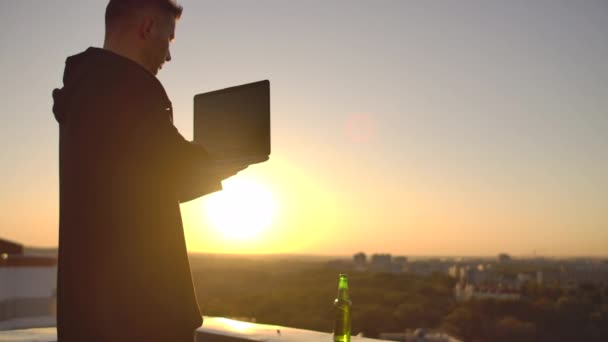 En programmerare med laptop sitter på taket och skriver kod vid solnedgången. Fjärrstyrd frilansare. Frihet att arbeta. Skriva på ett tangentbord vid solnedgången med utsikt över staden — Stockvideo