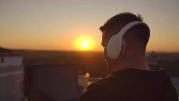 Hipster hombre con un portátil en el borde del techo. Freelancer trabajando. Internet móvil inalámbrico. Trabaja en Internet con auriculares y escuchando música . — Vídeo de stock