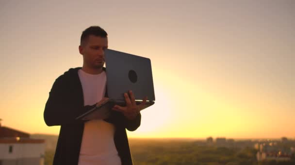 Ein Mann mit Laptop steht bei Sonnenuntergang auf dem Dach des Gebäudes und schreibt einen Code auf die Tastatur. Hacker mit Laptop — Stockvideo