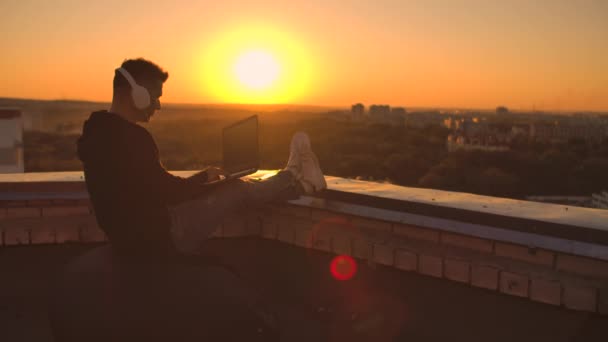 En programmerare med bärbar dator i hörlurar sitter på taket och skriver kod vid solnedgången och lyssnar på musik. Fjärrstyrd frilansare. Frihet att arbeta, Skriva på tangentbord vid solnedgången med utsikt över staden — Stockvideo