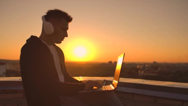 Чоловік у светрі сидить на даху і слухає музику з навушниками, що друкуються на клавіатурі ноутбука . — стокове відео