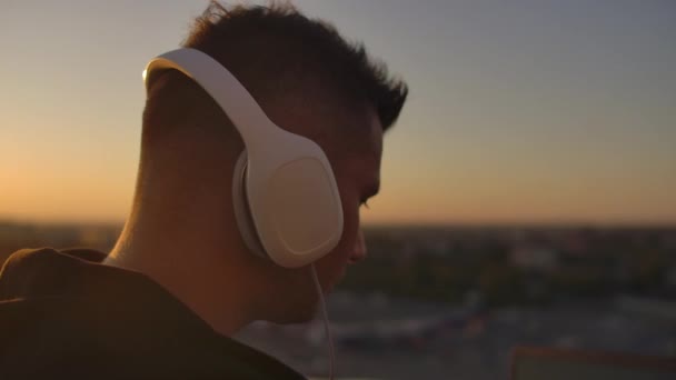 Крупним планом чоловік в навушниках слухає музику на даху під час заходу сонця з видом на місто . — стокове відео