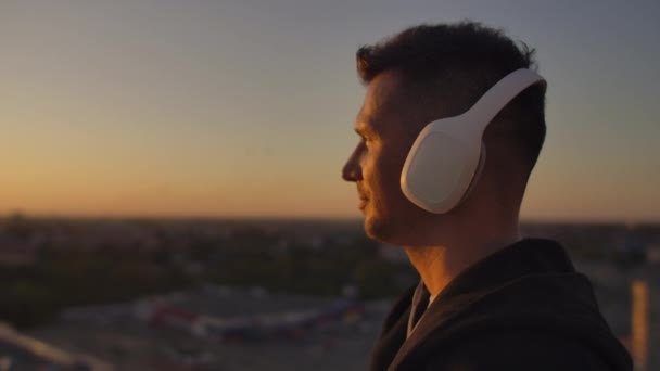 Крупним планом чоловік в навушниках дивиться на місто з висоти хмарочоса на заході сонця. Розслабтеся, слухаючи музику. Насолоджуйтесь прекрасним видом на місто під час заходу сонця з даху з навушниками — стокове відео