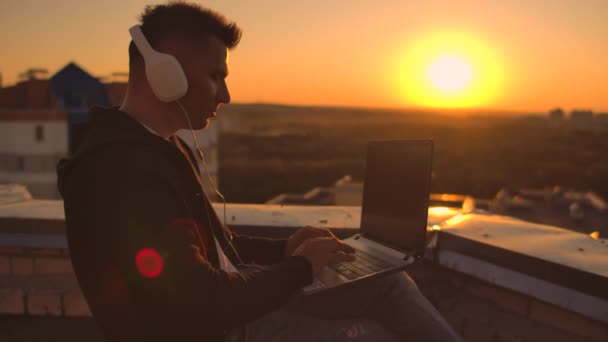 Um homem com capuz senta-se no telhado e ouve música com fones de ouvido digitando em um teclado de laptop . — Vídeo de Stock