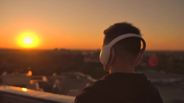 Un hombre camina en el techo al atardecer con auriculares mirando a la ciudad desde la altura de un rascacielos al atardecer. Relájese mientras escucha música . — Vídeos de Stock