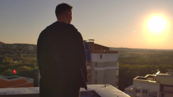 Um homem com um laptop caminha no telhado ao pôr do sol e olha para a cidade de uma altura ao pôr do sol — Vídeo de Stock