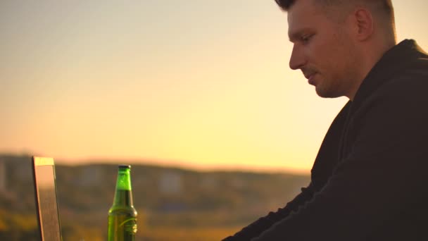 Joven hombre freelancer se sienta en el techo con un ordenador portátil y cerveza y tipos en el teclado — Vídeo de stock