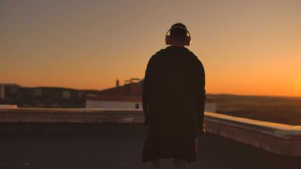 Um homem caminha no telhado ao pôr-do-sol com fones de ouvido olhando para a cidade da altura de um arranha-céu ao pôr-do-sol. Relaxe enquanto ouve música . — Vídeo de Stock