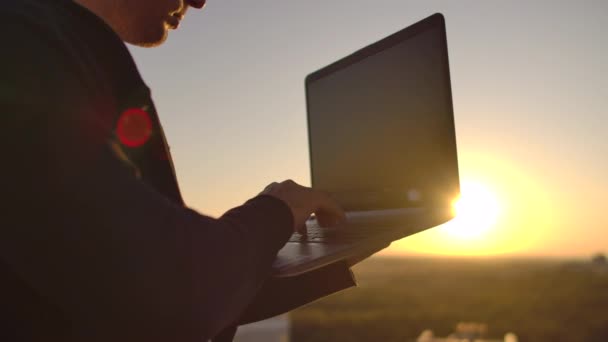 Un programador con un portátil se sienta en el techo y escribe código al atardecer. Trabajo remoto freelancer. Libertad para trabajar. Escribir en un teclado al atardecer con una vista de la ciudad — Vídeo de stock