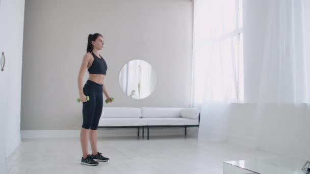 Žena zvedne paže s činkami a cvičí ramena ve svém světle bílém bytě proti pohovce a oknu — Stock video