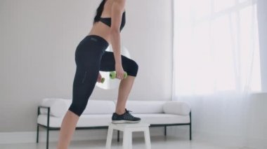 Genç, esmer bir kadın, elinde dambıllarla, parlak bir iç dairede, koltuğun üzerinde volta atıyor.