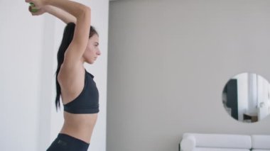 Genç bir kız, triseps egzersizi yaparken kollarını başının arkasından dambıllarla kaldırıyor. Kol kasları eğitimi