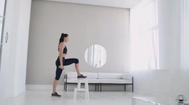 Genç, esmer bir kadın, elinde dambıllarla, parlak bir iç dairede, koltuğun üzerinde volta atıyor.