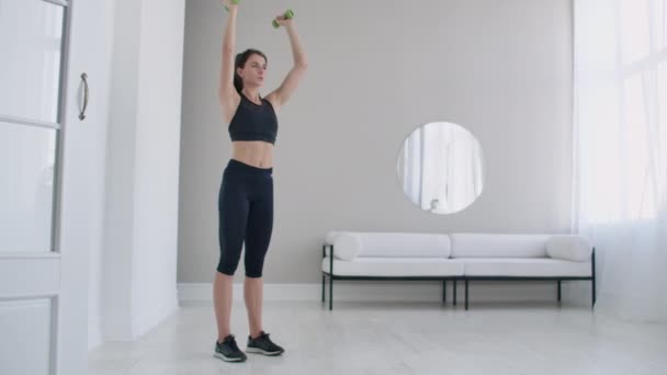 Una joven mujer caucásica levanta pesas para ejercicios de brazos y hombros. Levanta pesas sobre tu cabeza. — Vídeo de stock