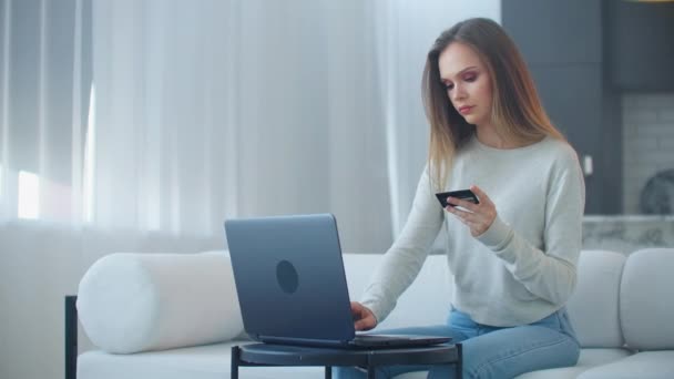 若い美しいブロンドの女性はノートパソコンの画面を見て、彼女の手に銀行カードを保持します。インターネット経由でリモートで銀行サービスを使用する — ストック動画