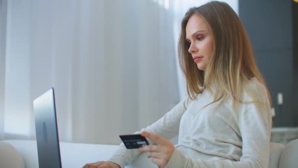 Ładna dziewczyna robi płatności online posiadanie karty bankowej przy użyciu nowoczesnego laptopa w domu siedzi na kanapie i uśmiechnięty. Finanse, zakupy w Internecie i koncepcja młodzieżowa — Wideo stockowe
