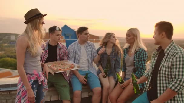 La compañía de seis jóvenes se comunican entre sí en el techo con pizza y cerveza. Las niñas miran a su alrededor al atardecer de verano . — Vídeos de Stock