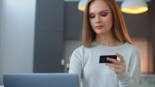 Chica bonita está haciendo el pago en línea celebración de la tarjeta bancaria utilizando un ordenador portátil moderno en casa sentado en el sofá y sonriendo. Finanzas, compras en internet y concepto juvenil — Vídeos de Stock