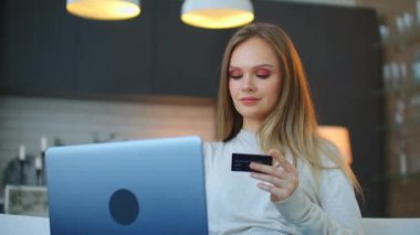 25 yaşında güzel bir kadın kucağında dizüstü bilgisayarla kanepede oturuyor ve online ödeme için kredi kartı kullanarak online bir mağazada seçilen ürünlerin parasını ödüyor.