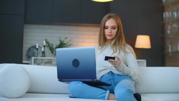 Menina bonita está fazendo pagamento on-line segurando cartão bancário usando laptop moderno em casa sentado no sofá e sorrindo. Finanças, compras na internet e conceito de juventude — Vídeo de Stock