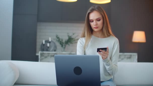 Blondynka w swetrze siedzi w domu na kanapie z laptopem patrzącym na ekran, trzymająca kartę kredytową i pisząca na klawiaturze. — Wideo stockowe
