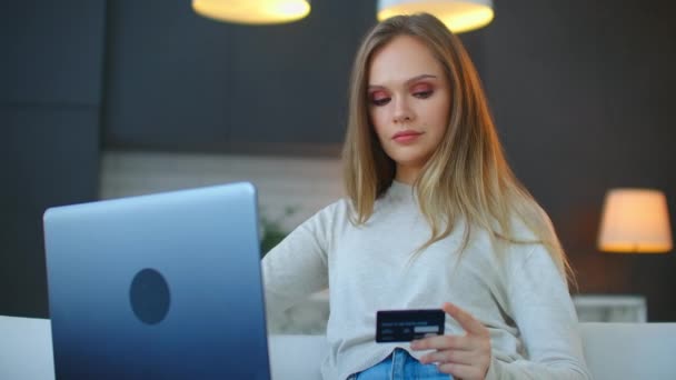 Egy gyönyörű szőke ül a kanapén egy laptoppal és egy hitelkártyával tanulmányozza a hiteltörténetét egy online bankban. — Stock videók