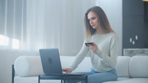 セーターのブロンドはソファの上に家に座っているラップトップを使用してインターネット経由で簡単に支払いを行います — ストック動画