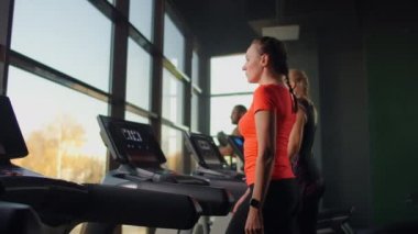 Büyük bir panoramik pencerenin yanında koşu bandında yürüyen bir grup insan. Grup kardiyo çalışması. Fitness derslerine başlıyorum. Genç, güzel kadınlar ve erkekler spor giyim odasında.