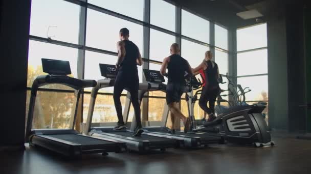 Un grupo de personas corriendo en una cinta de correr en una sala de fitness realizando un ejercicio cardiovascular. Hombres y mujeres entrenan juntos Correr en interiores, calentar antes del entrenamiento en cámara lenta — Vídeo de stock