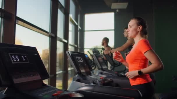 Uma jovem mulher bonita e três pessoas correndo em uma esteira em uma sala de fitness realizando um treino de cardio. Corrente interior, aquecimento antes do treino em câmara lenta — Vídeo de Stock