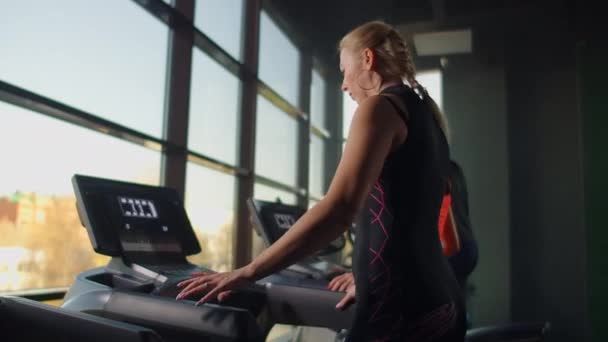 Une femme s'approche du tapis roulant et l'allume pour commencer l'entraînement. Inclure un tapis roulant dans la salle de fitness — Video