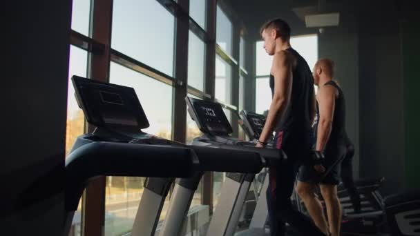 Män går på löpband i fitnessrummet. En grupp människor går på löpband nära ett stort panoramafönster. Inledande av träningspass. Unga vackra kvinnor och män i sportkläder — Stockvideo