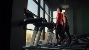 Büyük bir panoramik pencerenin yanında koşu bandında yürüyen bir grup insan. Grup kardiyo çalışması. Fitness derslerine başlıyorum. Genç, güzel kadınlar ve erkekler spor giyim odasında.