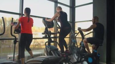 Genç, formda bir kadın, spor salonunda eliptik bir eğitmen kullanıyor. Bir grup genç kadın spor salonunda spor malzemeleriyle antrenman yapıyor. Kamerayı sabit tut.