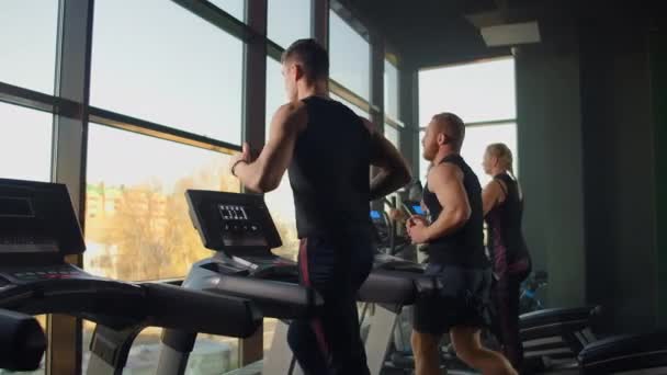 Dois homens correndo em uma esteira no fundo de uma grande janela na sala de fitness. Jovens a correr na sala de fitness. Exercícios aeróbicos no ginásio — Vídeo de Stock