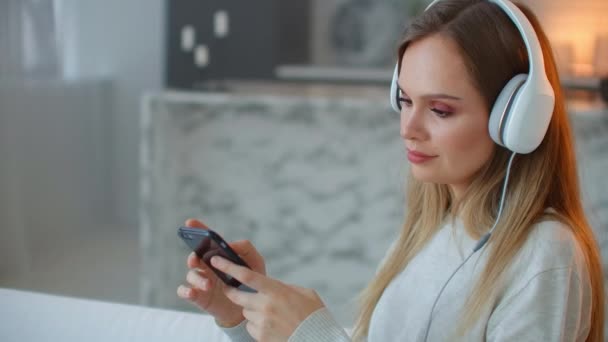 Glücklicher amerikanischer Teenager, der es sich zu Hause auf dem Sofa mit dem Smartphone gemütlich macht. junge Frau streamt nachts Musik auf Mobiltelefon und hört mit drahtlosen Kopfhörern — Stockvideo